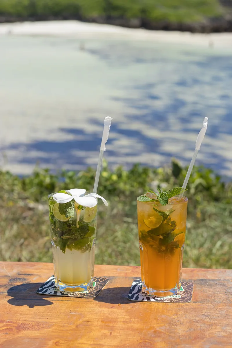 Two tall cocktails on a wooden table by the water