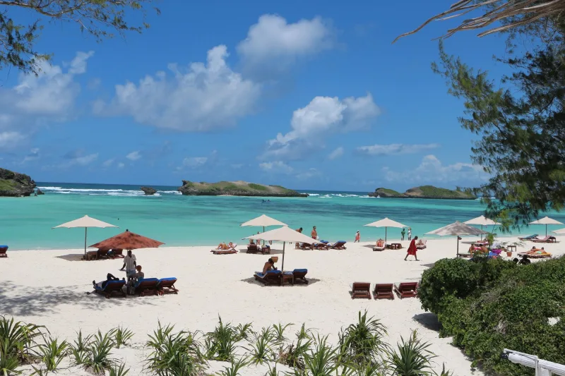 Guests on loungers under umbrellas along a wide stretch of white sand, with a Maasai askari in red walking the beach and green islets out in the bay.