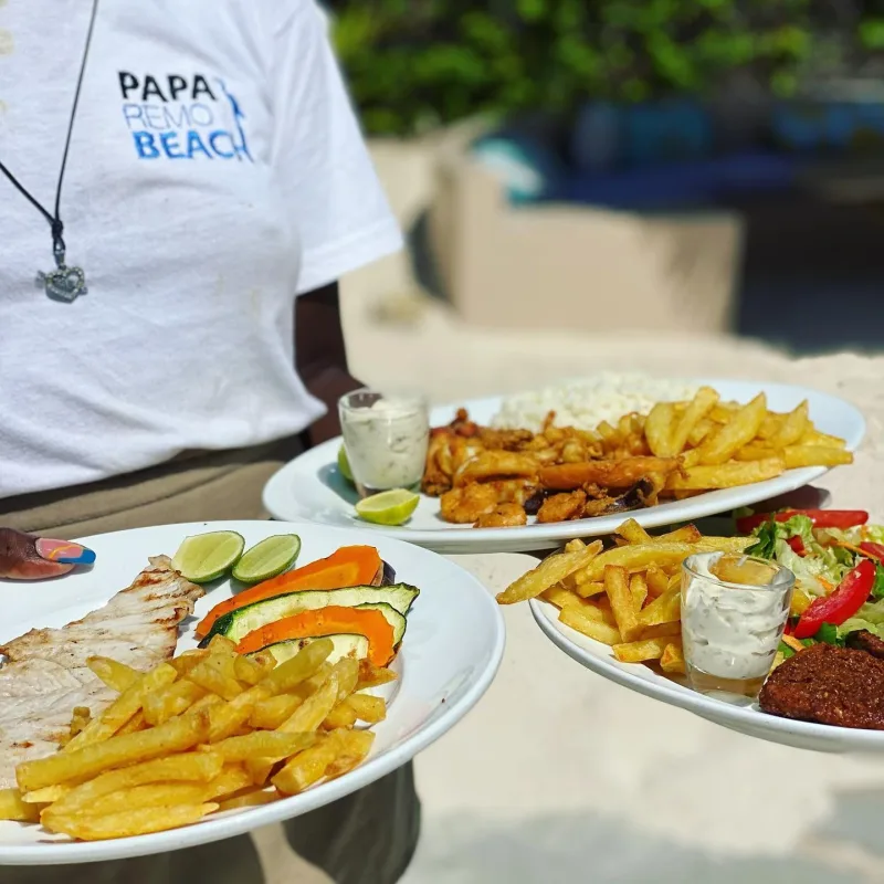 A member of staff in a Papa Remo Beach shirt carrying three plates — grilled fish with vegetables and chips, fried calamari with rice, and a salad.