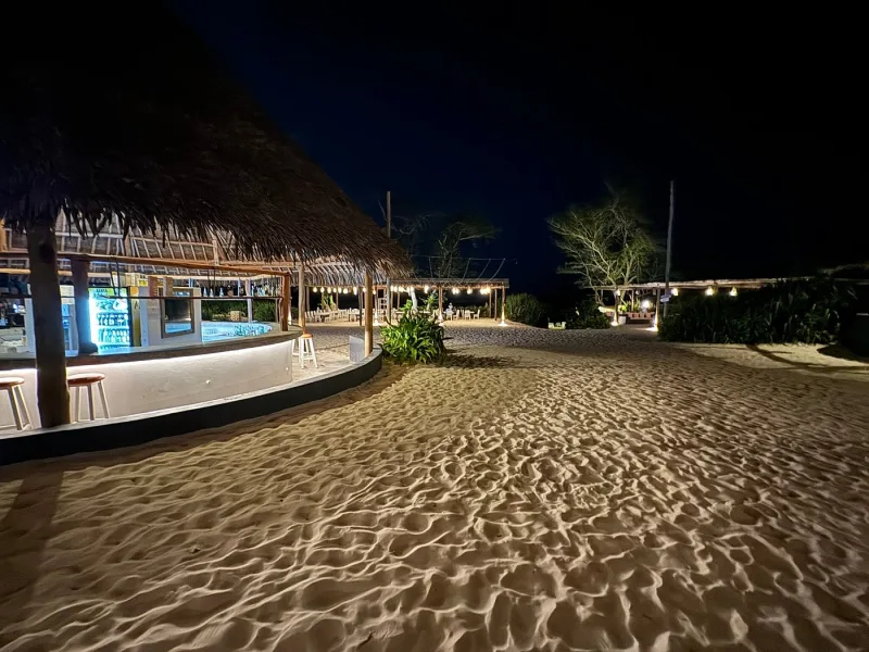 The curved beach bar lit under its makuti roof at night, with stools at the counter and lit tables further along the sand.