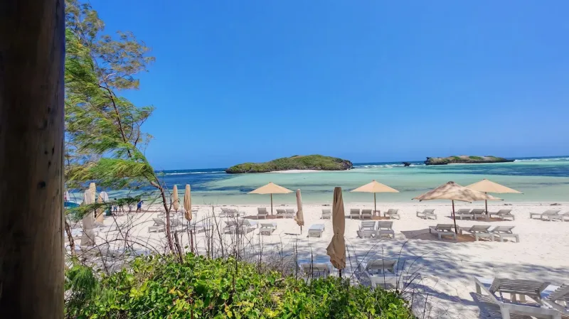 Umbrellas and loungers on white sand seen from among the trees, with the turquoise lagoon and two islets beyond.