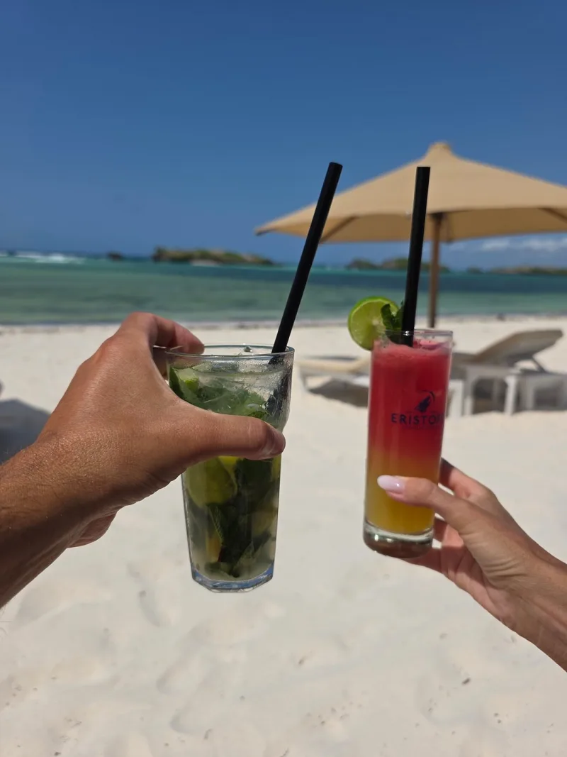 Two hands touching a mojito and a layered red and orange cocktail together, with the lagoon and a cream umbrella behind.