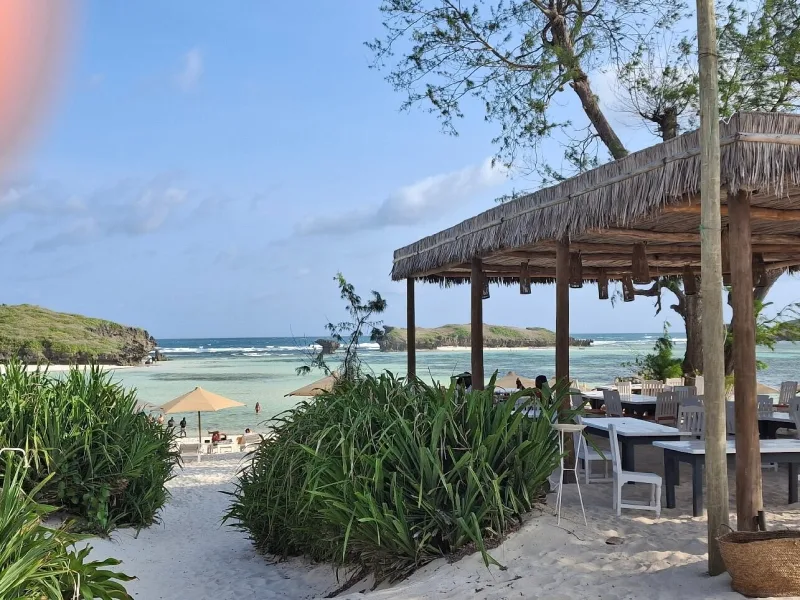 The makuti-roofed restaurant with its white tables, seen from the sandy path down to the beach, with the lagoon and islets beyond.