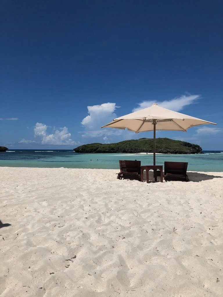 A cream umbrella shading two wooden loungers on empty white sand, with a green islet across the turquoise water.