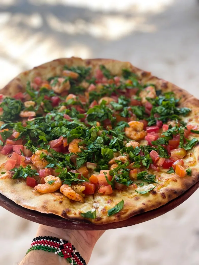 A thin, wood-fired pizza topped with prawns, chopped tomato and shredded basil, held on a wooden board.