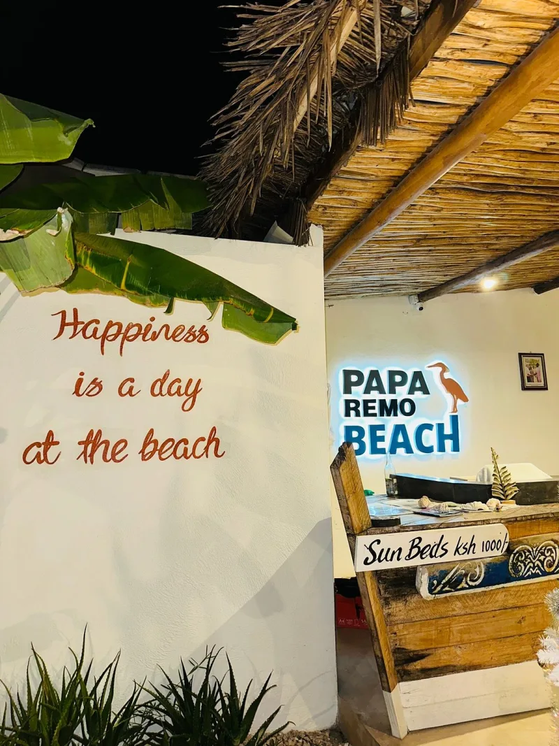 A whitewashed wall painted with the words "Happiness is a day at the beach", beside a reception desk under a makuti roof with the Papa Remo Beach sign lit above it.
