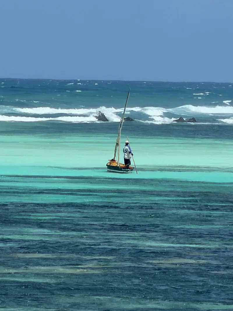 A man standing at the stern of a small dhow, poling it across the turquoise lagoon, with surf breaking on the reef behind.