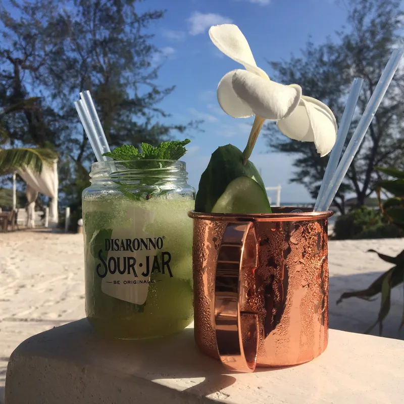 A mint cocktail in a glass jar beside a copper mug garnished with cucumber and a frangipani flower, on a white ledge above the sand.