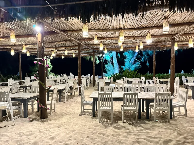 Tables and chairs set out on the sand under a makuti roof hung with woven lanterns at night, with a tree lit blue behind.
