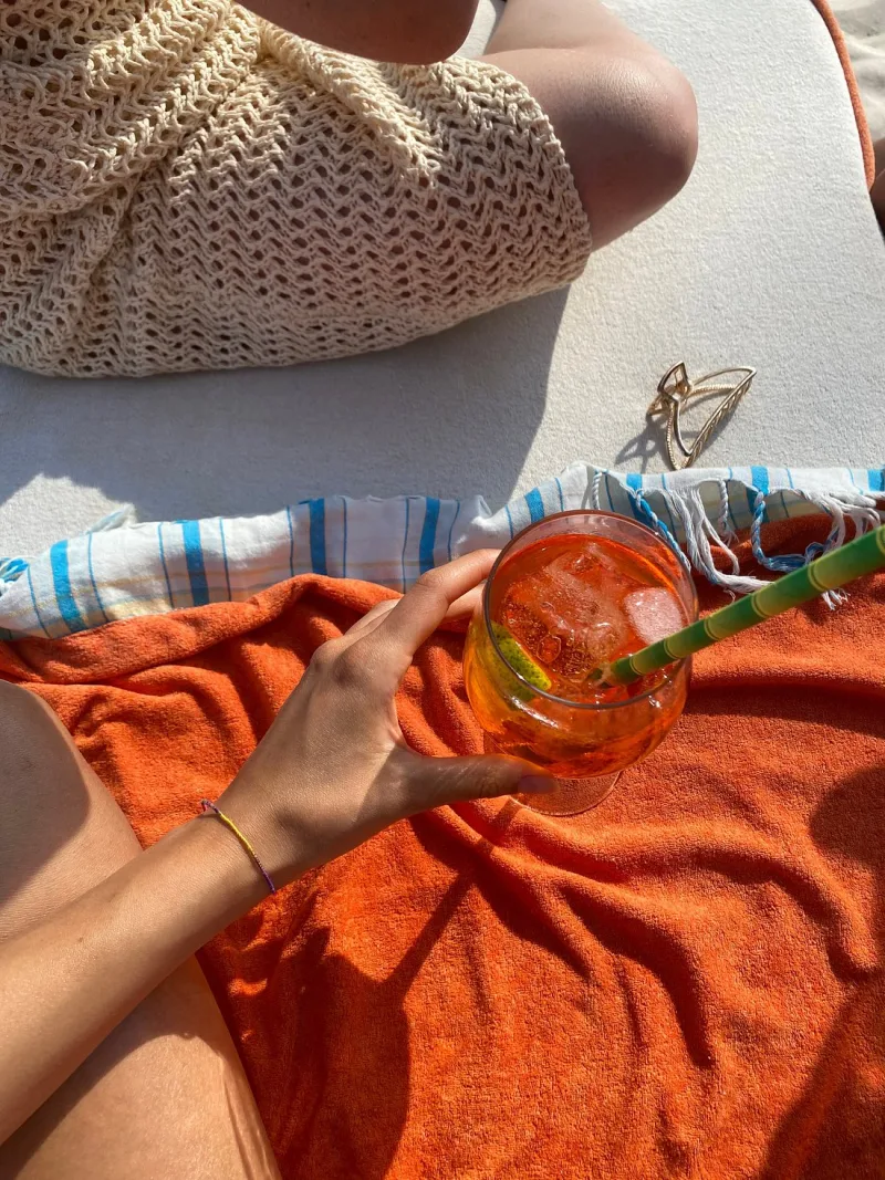 A person resting on a sun lounger with an orange towel, holding a tall iced orange cocktail with a green straw.